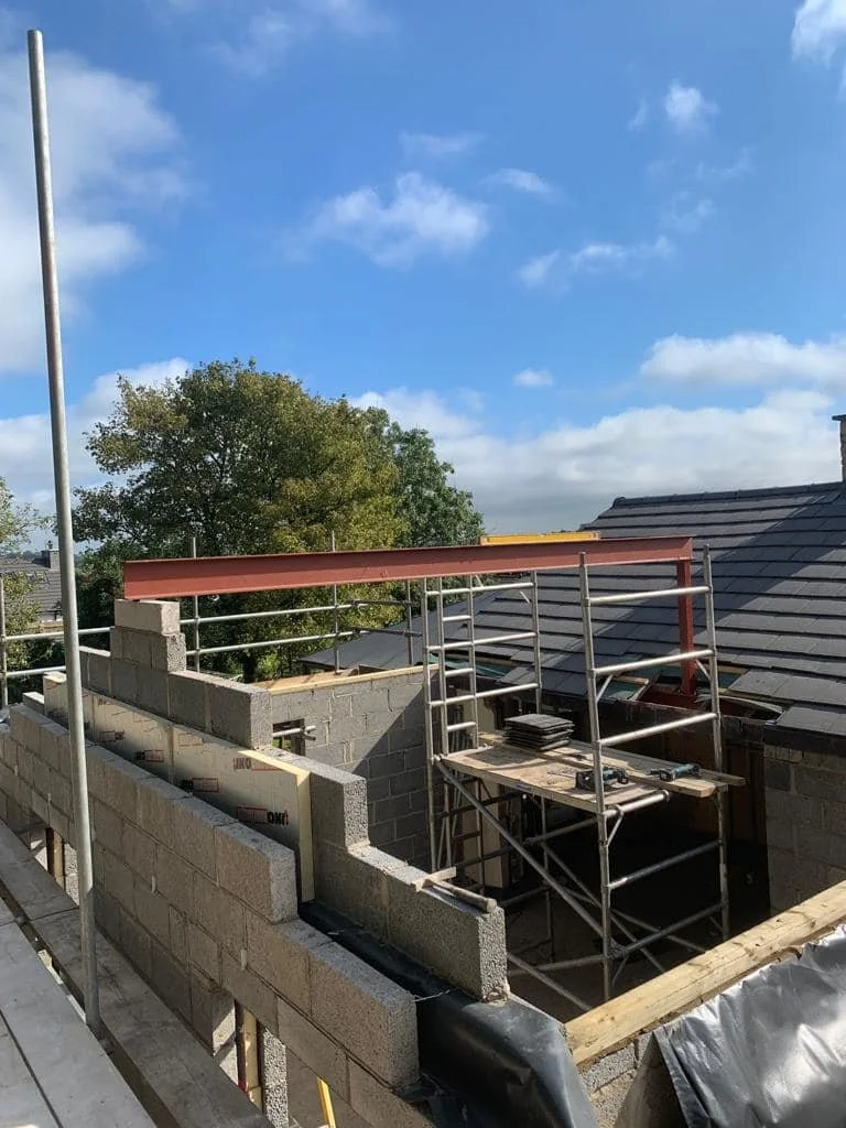 Construction site with a partially built cinder block wall, scaffolding, and framing for a new structure under a partly cloudy sky.