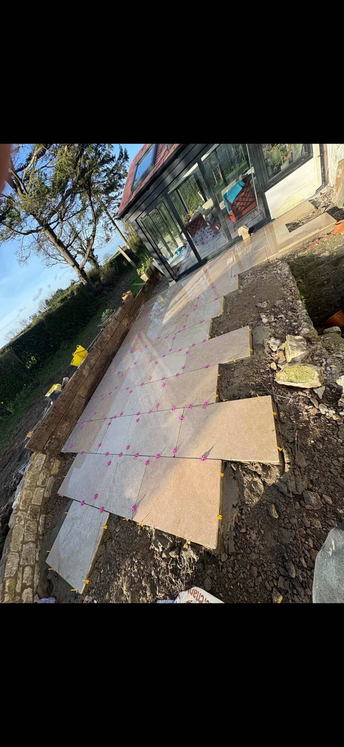 Unfinished outdoor patio with stone tiles being installed, construction tools and materials scattered around, and a glass-enclosed house in the background.