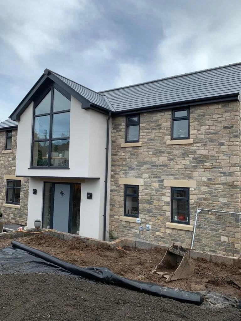 A modern, multi-story house with a stone facade and large glass windows, located in a residential area, with an ongoing landscaping project in the front yard.