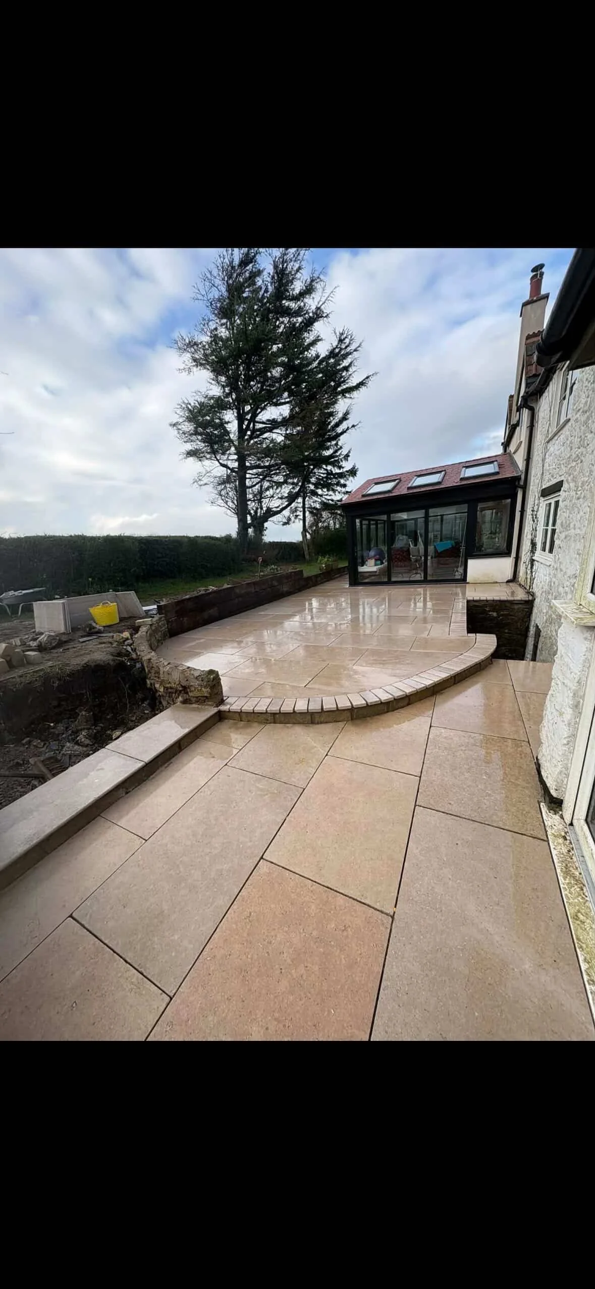 Image of a newly constructed patio with beige stone tiles, partially wet from recent rain, next to a house with a conservatory and large window, a tall tree in the background under a cloudy sky.
