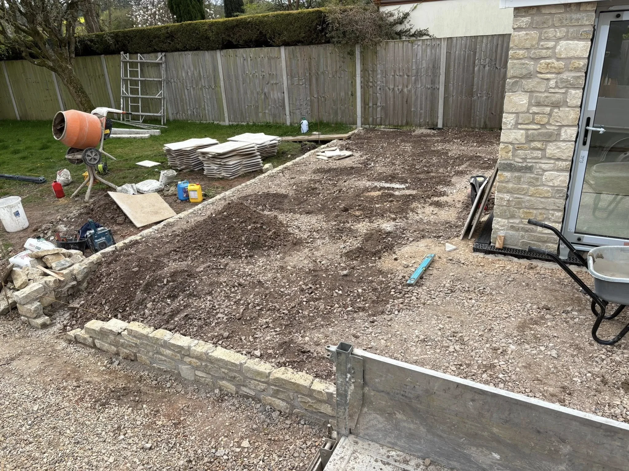 Backyard construction site with dirt and gravel, construction tools, wheelbarrow, stacked tiles, cement mixer, ladder, and building materials, surrounded by a wooden fence and a stone wall.