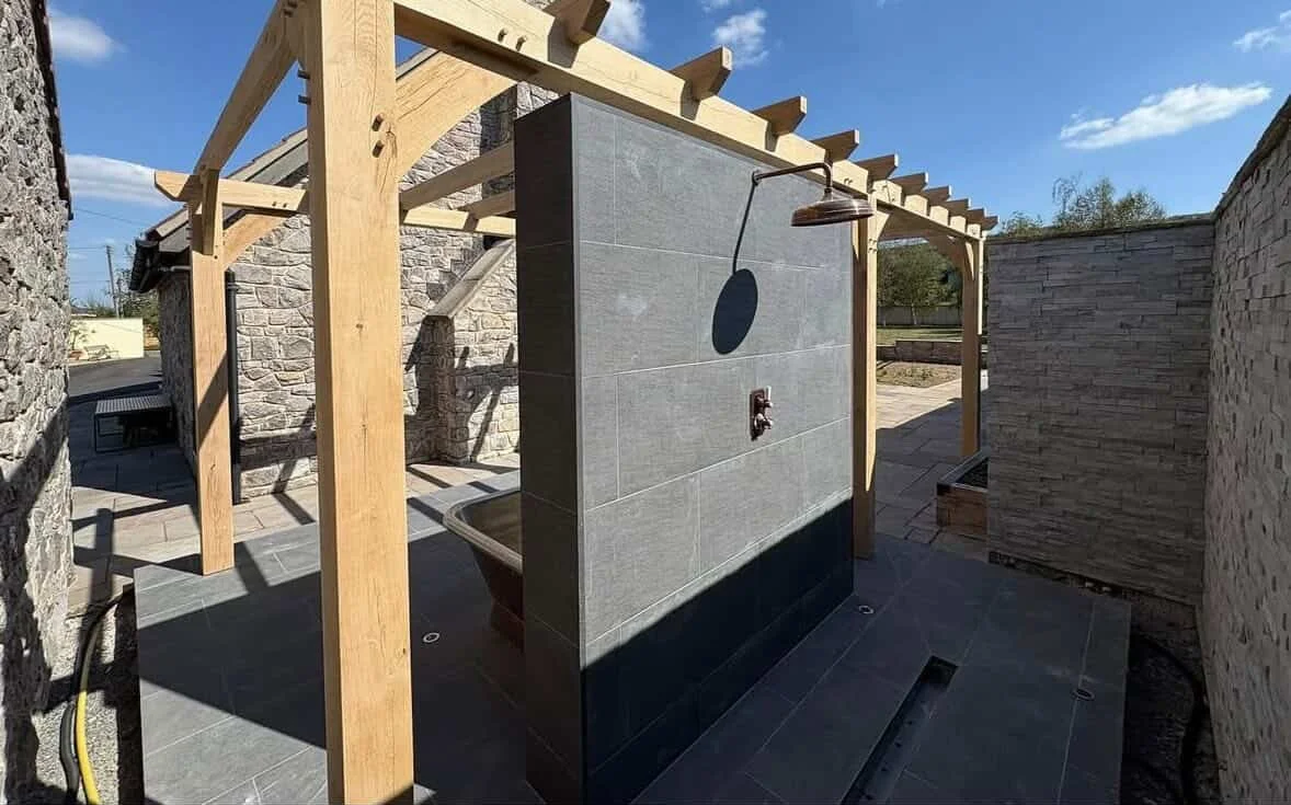 Outdoor shower with gray tiled wall, metal showerhead, and low black tiled base, under a wooden pergola, in a yard enclosed by stone walls.