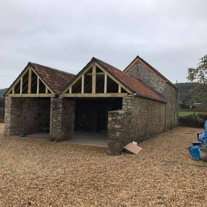 A stone barn with two wooden gabled roofs under construction, surrounded by gravel and construction materials, in a rural setting.