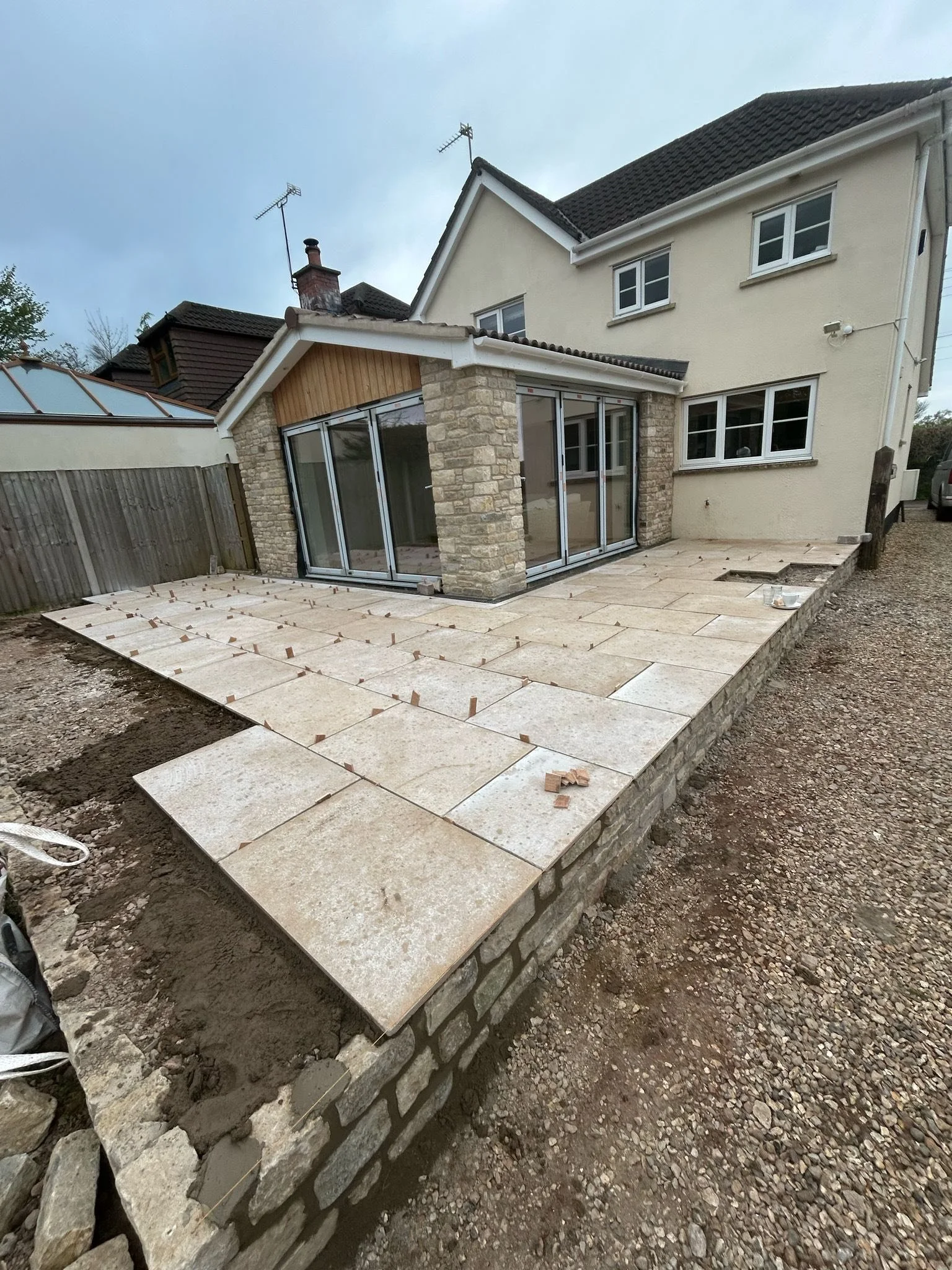 Construction of a new patio with stone tiles in the backyard of a house, partially completed, with some tiles still being installed and construction tools nearby.