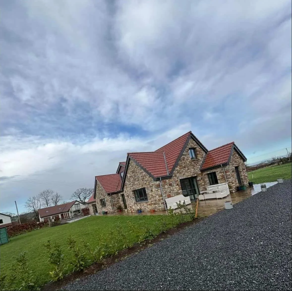 A stone house with red tile roof and black window frames, with a patio area, on a cloudy day with some rain, and a grassy yard surrounding the house.