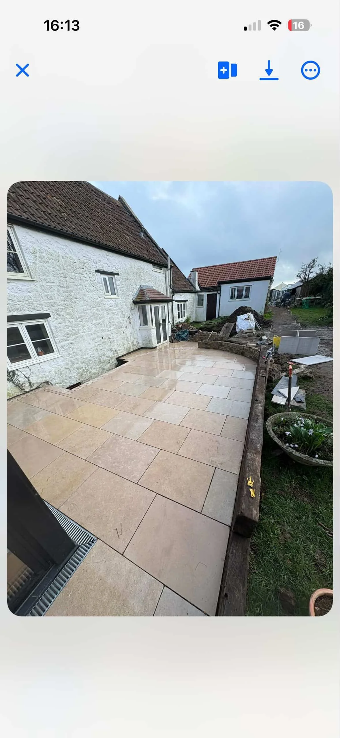 Freshly installed beige stone patio outside a white house with a red tile roof, surrounded by a garden area under construction with tools and building materials.