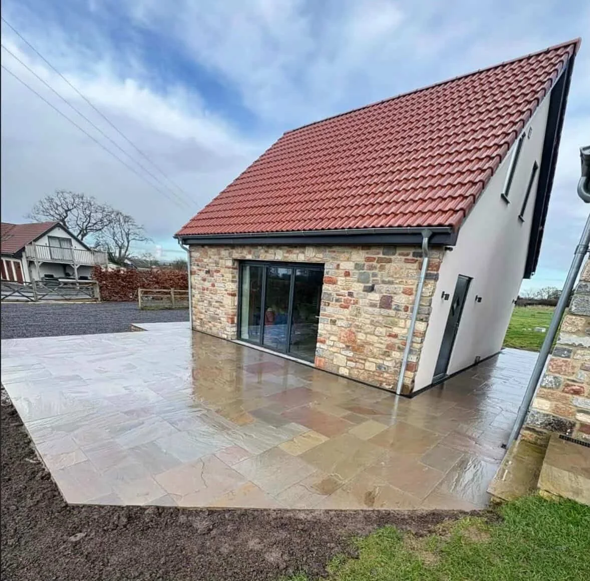 A house with a brick and stucco exterior, red tiled roof, and a large glass sliding door leading to a wet stone patio. The patio area is surrounded by a dirt border, and there are neighboring houses and trees in the background.