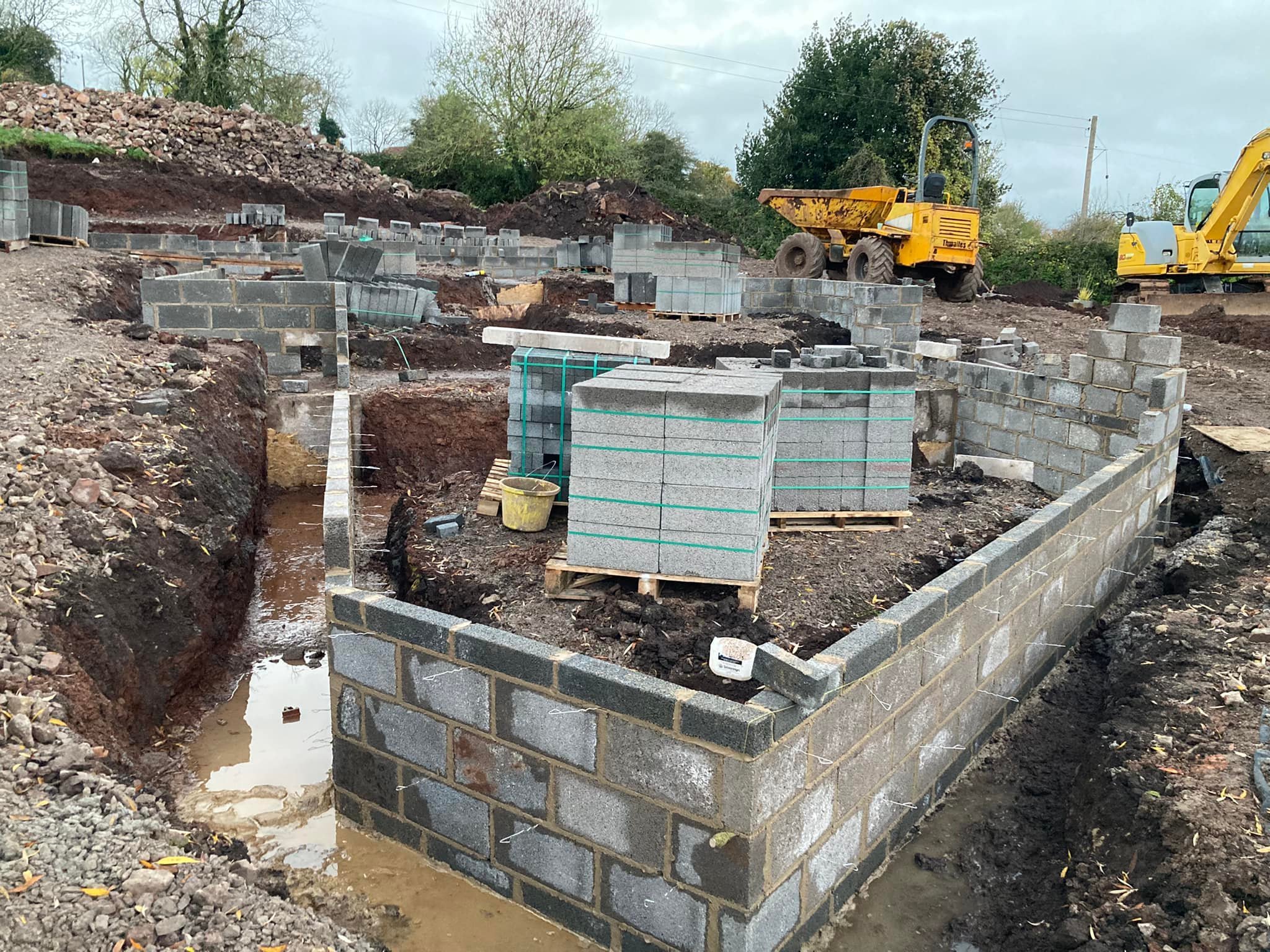 Construction site with partially built cinder block walls, construction materials, and construction vehicles, including a yellow dumper and excavator, in an outdoor setting.