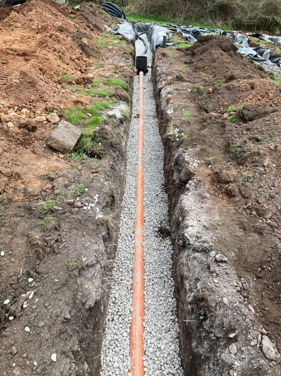 Construction site with a trench containing a long pipe, gravel, and black drainage pipes, surrounded by dirt and soil.