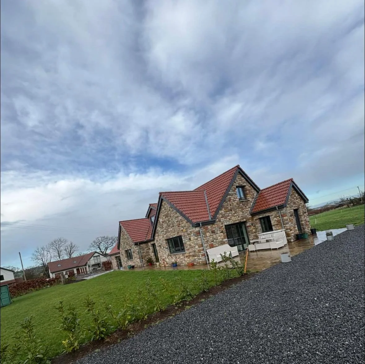 Stone house with red tile roofing on a cloudy day, with a green lawn and gravel path in front.