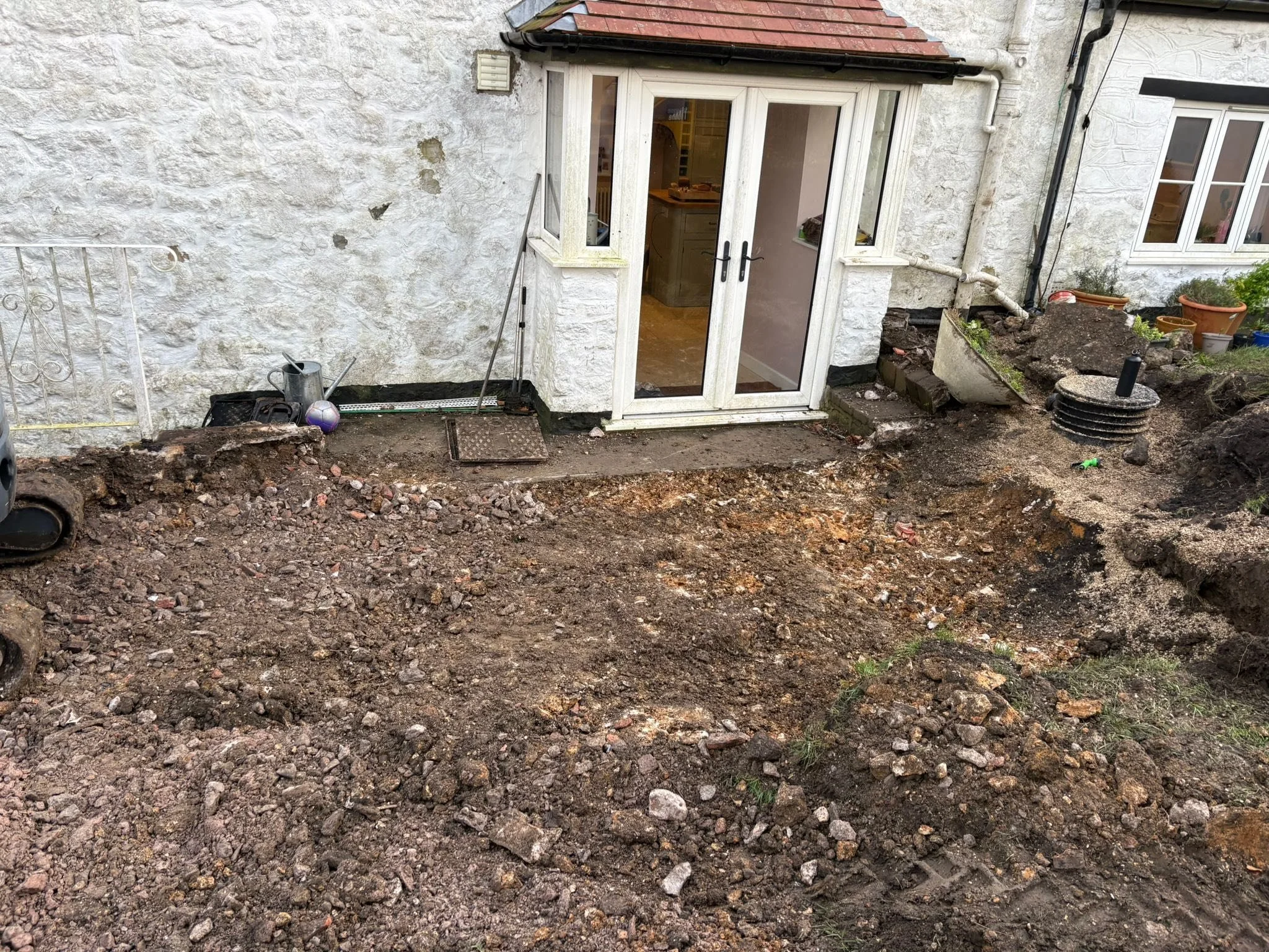 Backyard construction site with disturbed soil, tools, and a small porch with glass doors leading into a house with white exterior walls.
