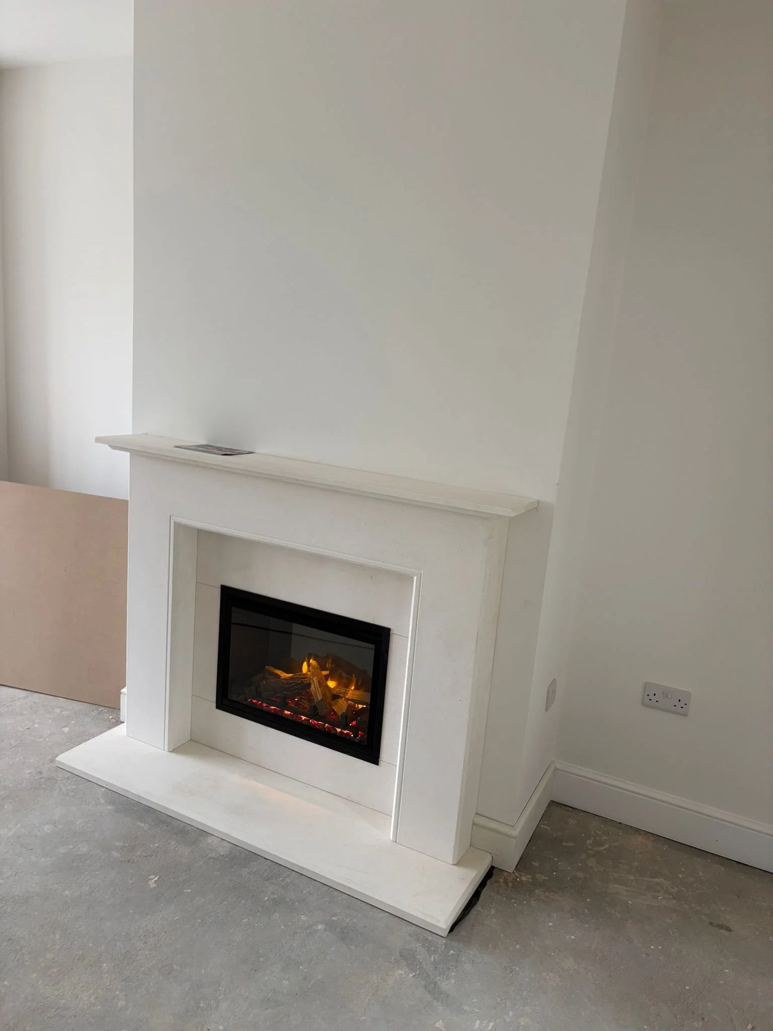 White fireplace with a decorative mantel and electric fire insert in a room with unfinished flooring and empty walls.