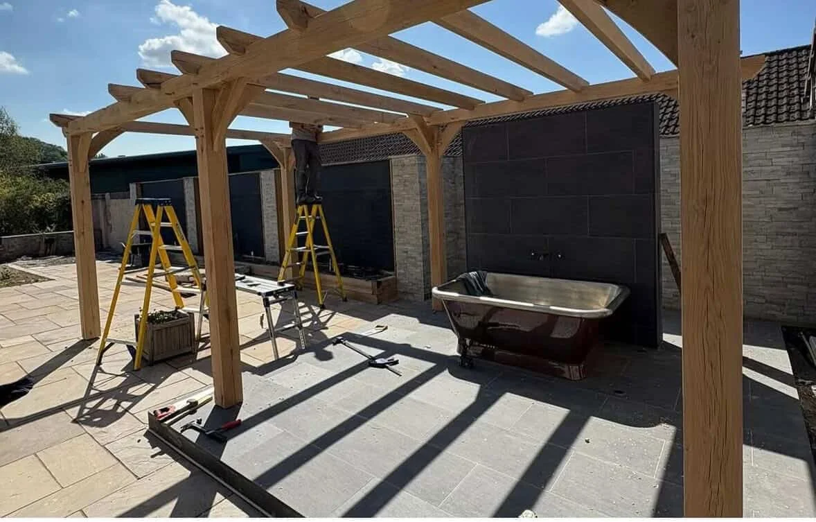Workers building a wooden pergola outdoors with ladders and tools, next to a black bathtub on a tiled patio.