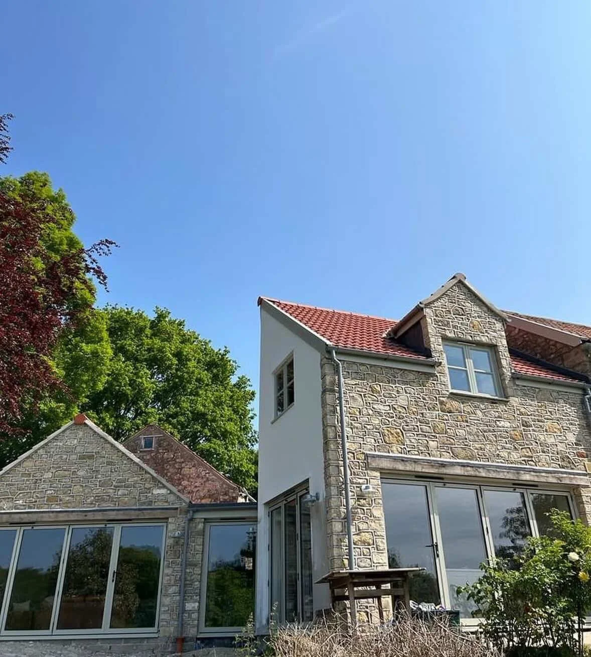 A stone and plaster house with large windows, a red tiled roof, and lush green trees under a clear blue sky.