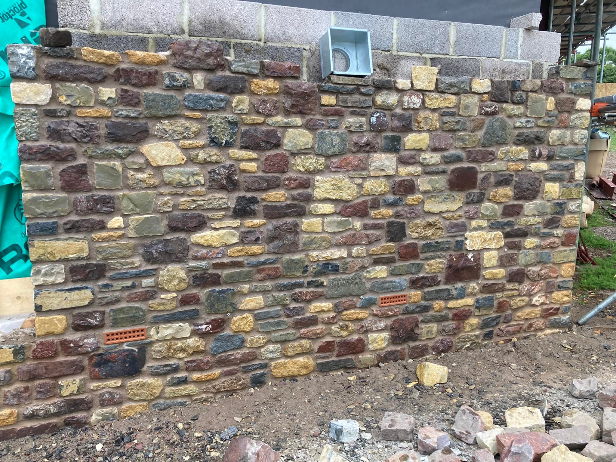A stone brick wall under construction with a mix of multicolored stones, visible at a construction site with loose rocks and dirt in front.