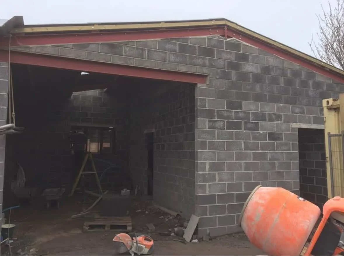Construction site of a brick structure with an unfinished roof and construction equipment in the foreground.