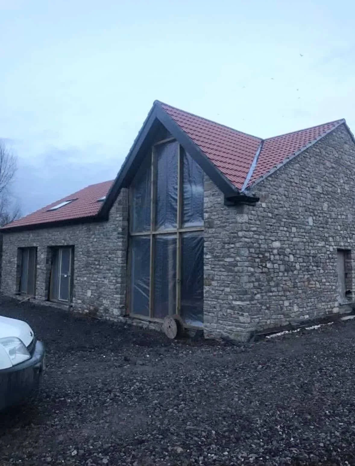 A stone house under construction with a steep red shingle roof, large front window covered with plastic, and a gravel driveway, during daytime.