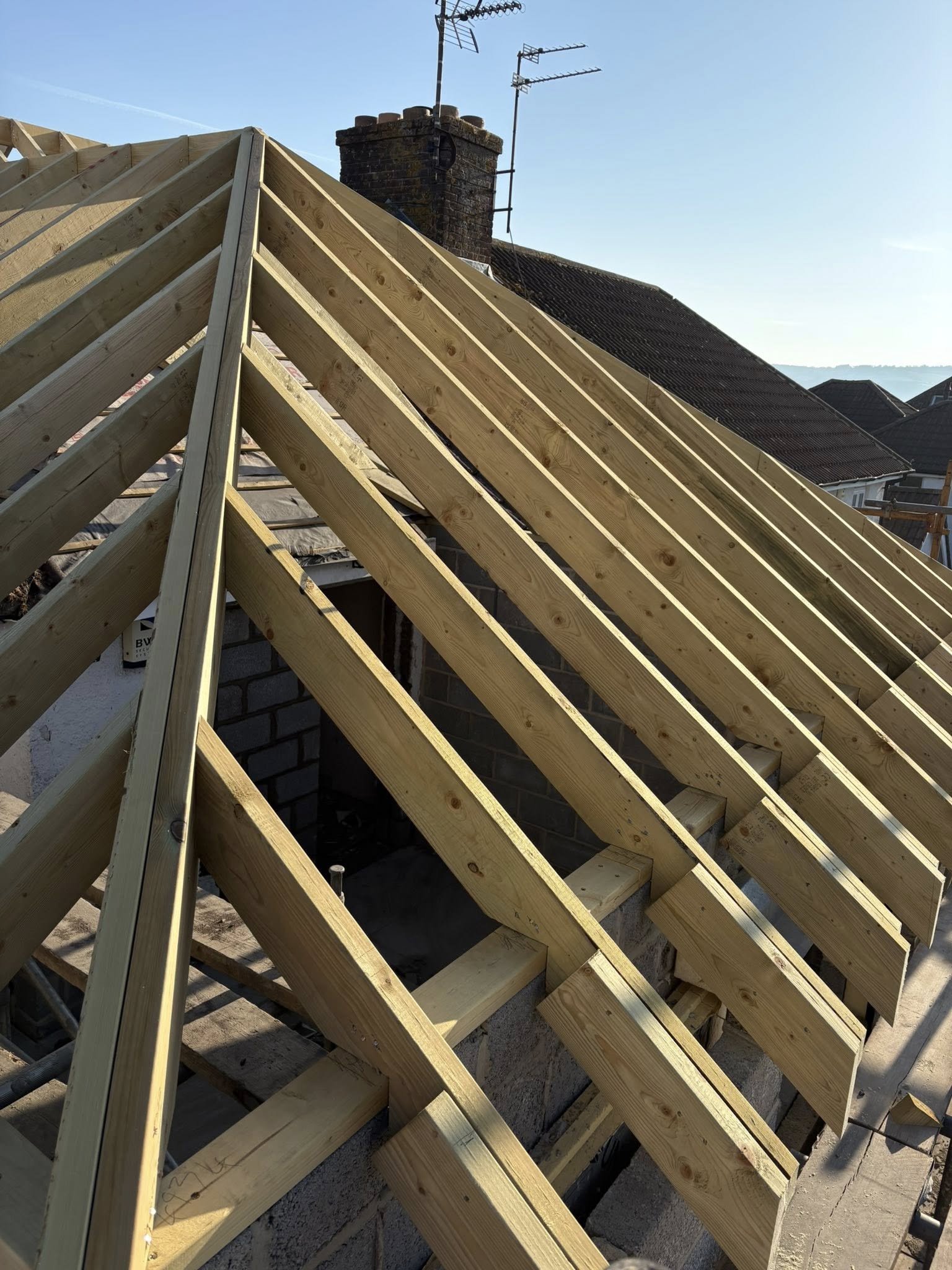 Construction site of a house roof with new wooden rafters installed, showing a chimney and TV antennas on neighboring roofs in the background.