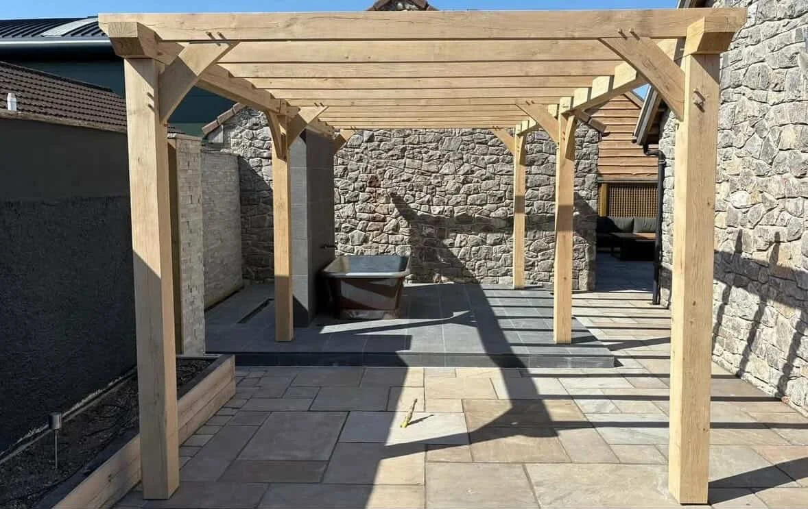 Wooden pergola over a paved backyard patio with stone wall, bathtub, and seating area.