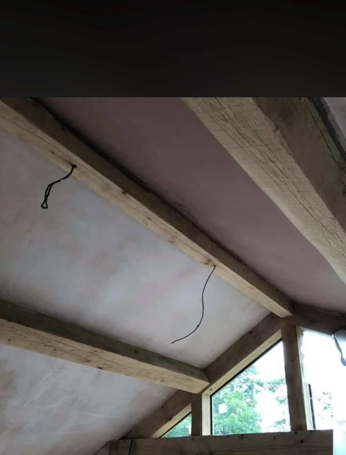 Interior view of a building under construction with exposed wooden beams, electrical wires hanging from the ceiling, and a window showing trees outside.