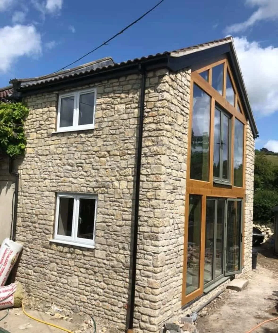 The image shows a two-story house with a stone exterior, several windows, and a large glass door framed with wood on an outdoor construction site.
