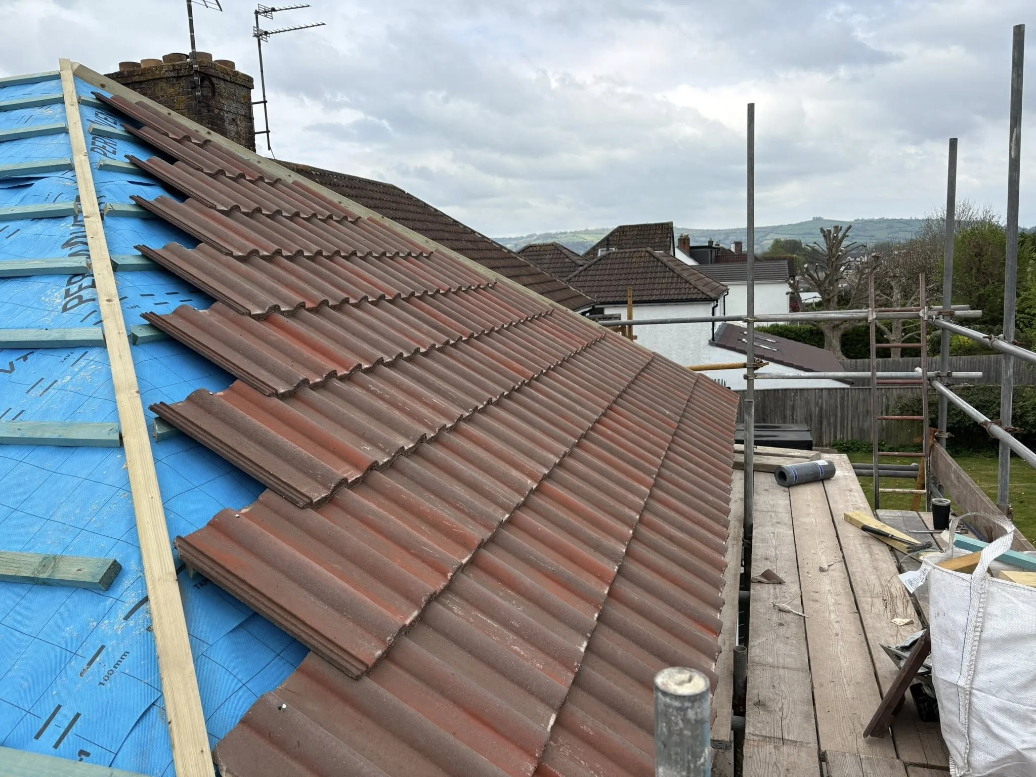 Underside of a roof under construction with partially installed brown roof tiles, blue underlayment, and scaffolding surrounding the building.