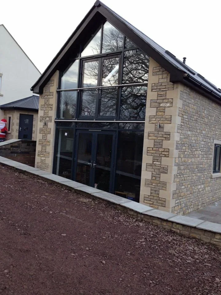 Modern house with a glass front, stone walls, and a triangular roof, situated on a sloped dirt yard.