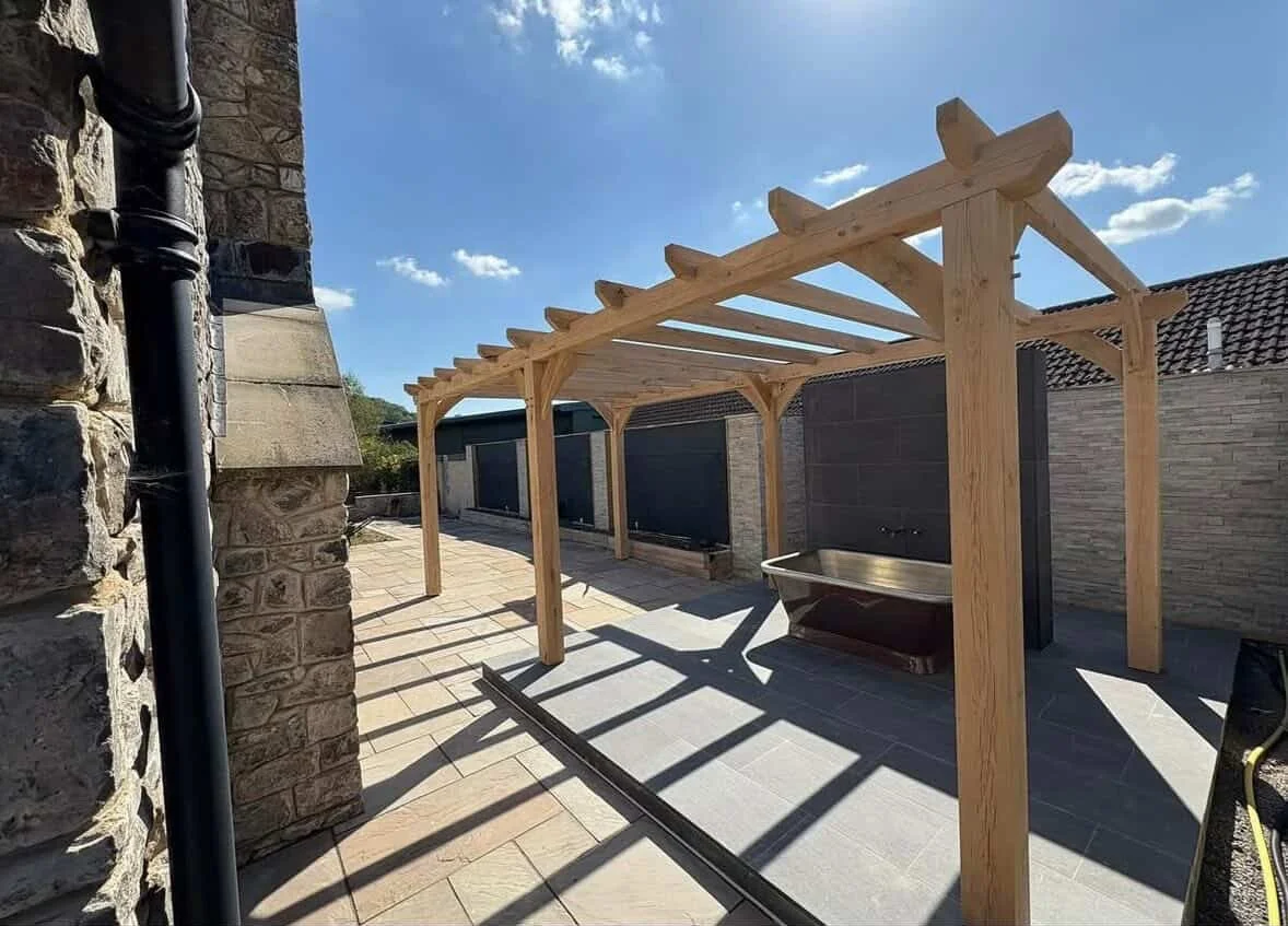 Wooden pergola structure under construction in an outdoor patio area with stone and brick walls.