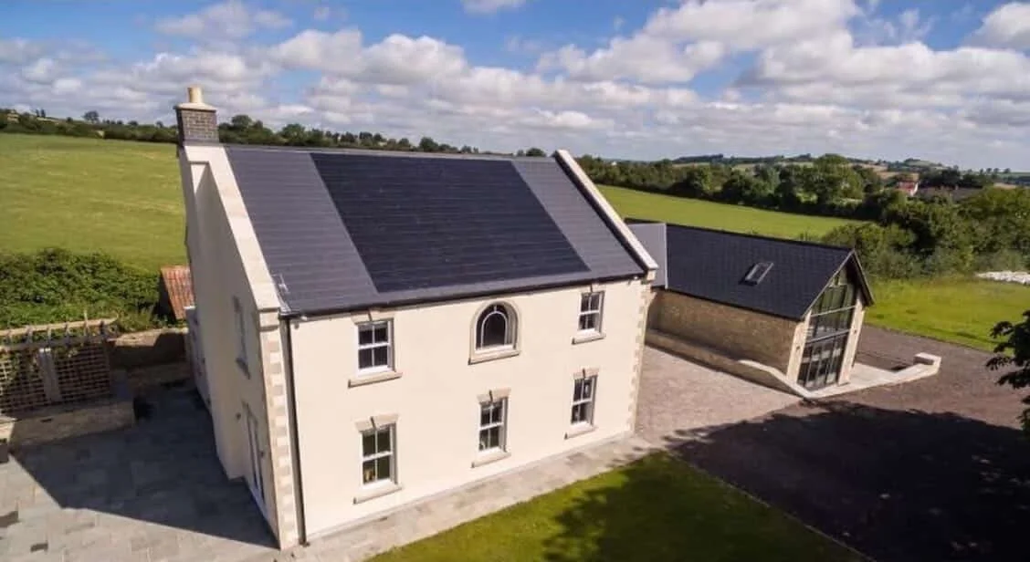 A white two-story house with a black roof, attached to a modern glass extension in a rural landscape with green fields and a partly cloudy sky.