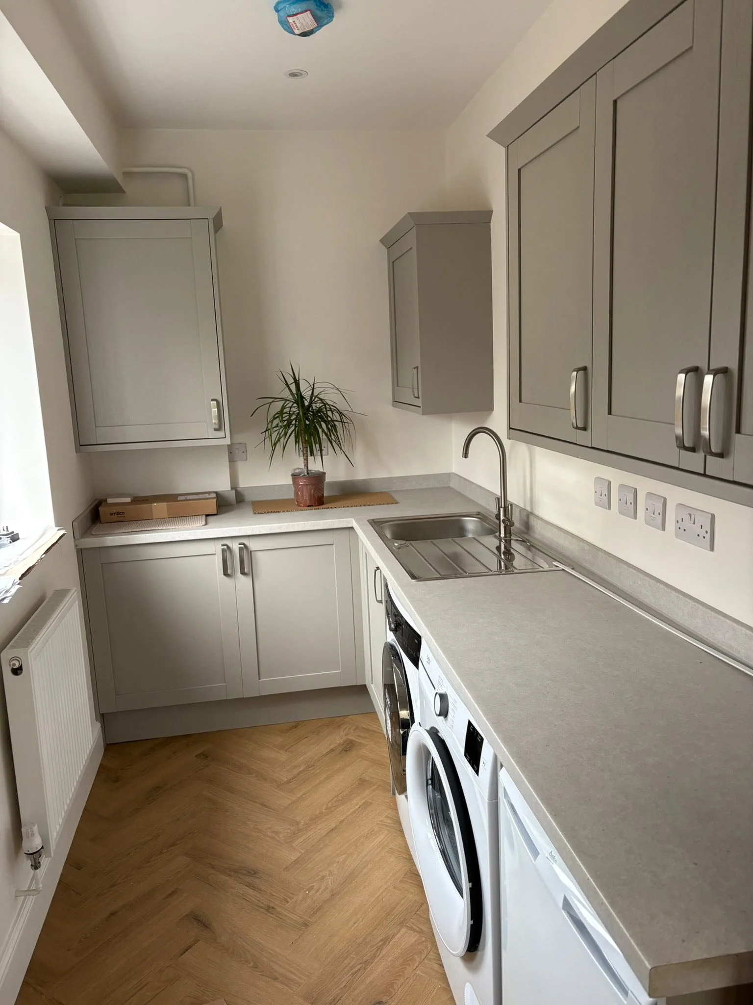 A small laundry room with gray cabinets, a gray countertop, a washing machine and dryer, a small sink, a window, a potted plant, and a ceiling light fixture.