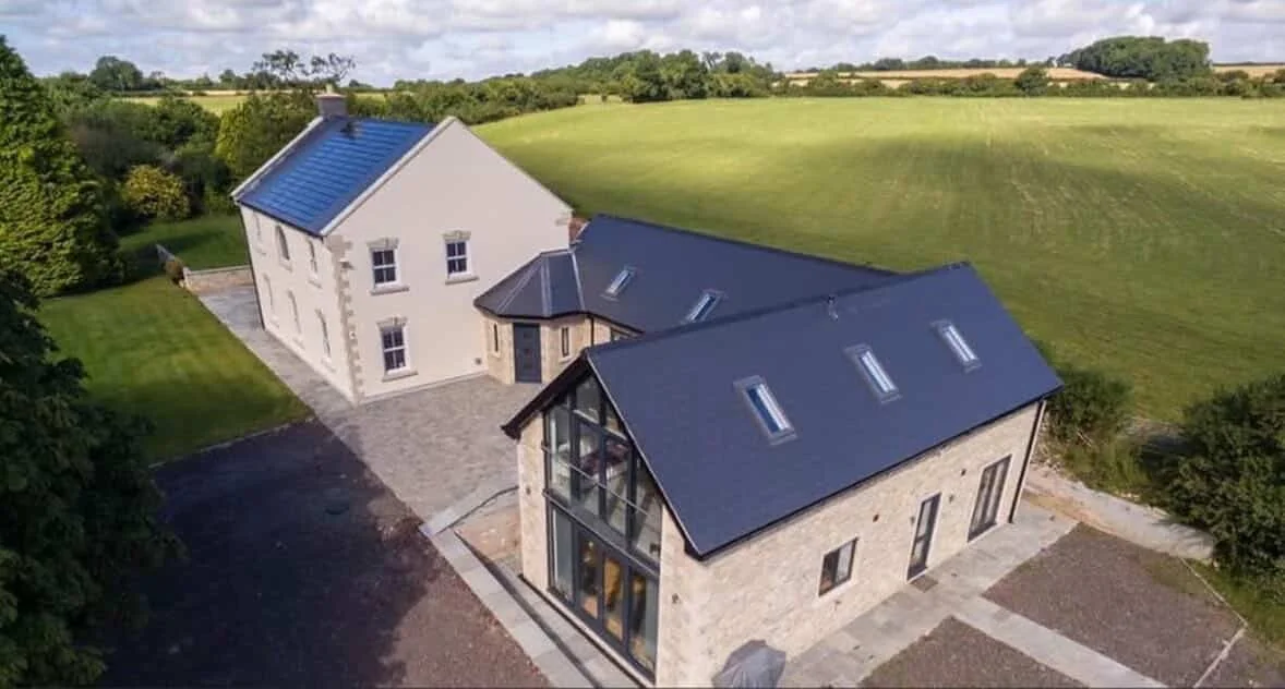 Aerial view of a modern two-story house with a stone exterior, large windows, and a dark metal roof, surrounded by green fields and trees.