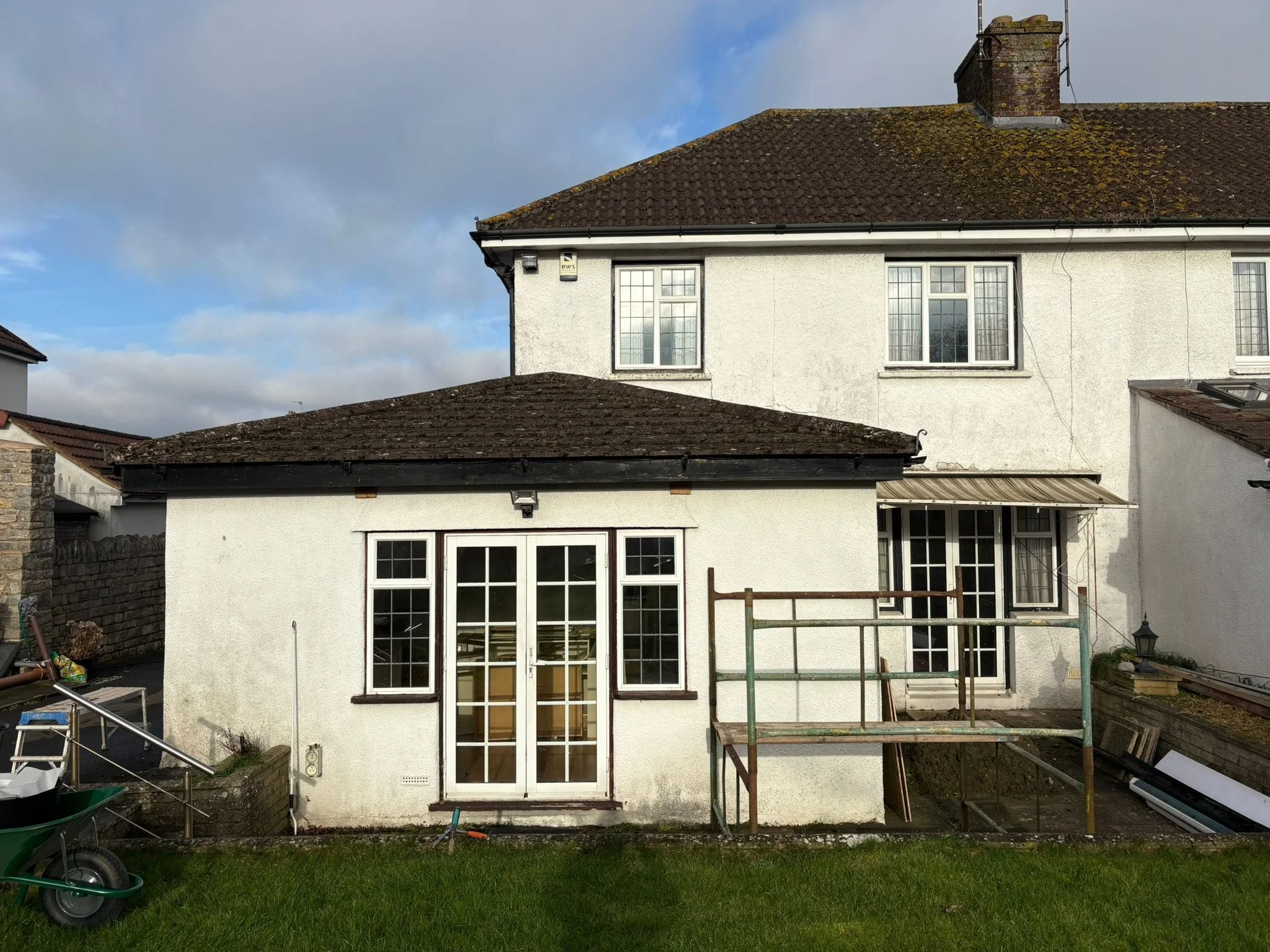 Two-story white house with a brown tiled roof, large windows, and a small extension with a glass door and windows. Scaffolding is set up outside, with construction tools and materials around, and a wheelbarrow in the yard.
