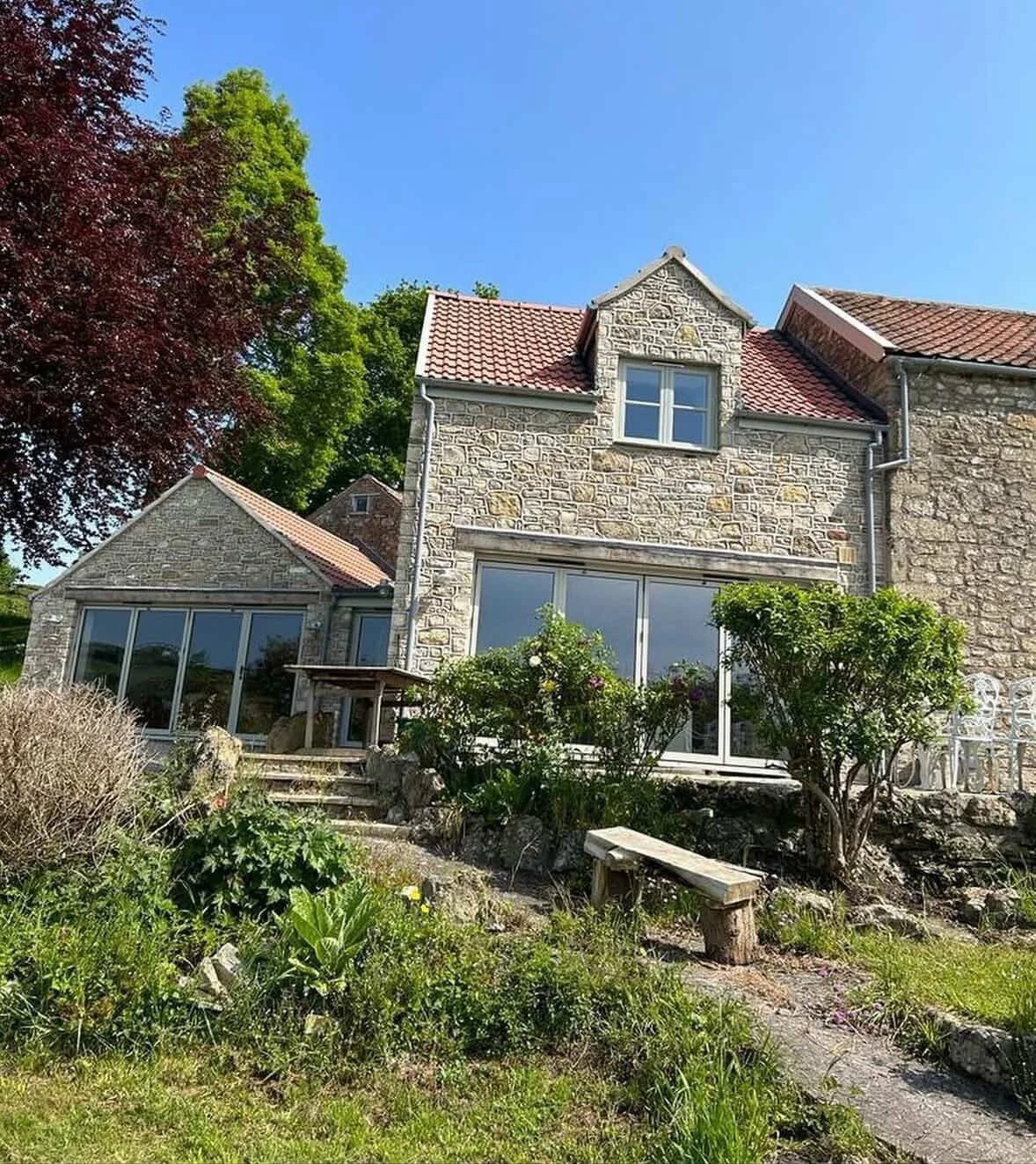 A stone house with large windows, a red-tiled roof, and green trees in the background, under a clear blue sky.