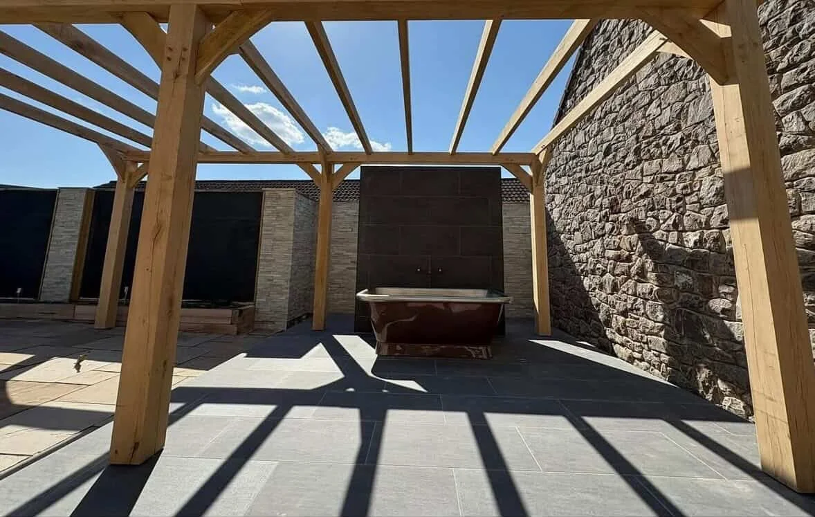 Wooden pergola frame on a tiled patio with brick and stone wall backgrounds, with a bathtub underneath, casting shadows on the ground.