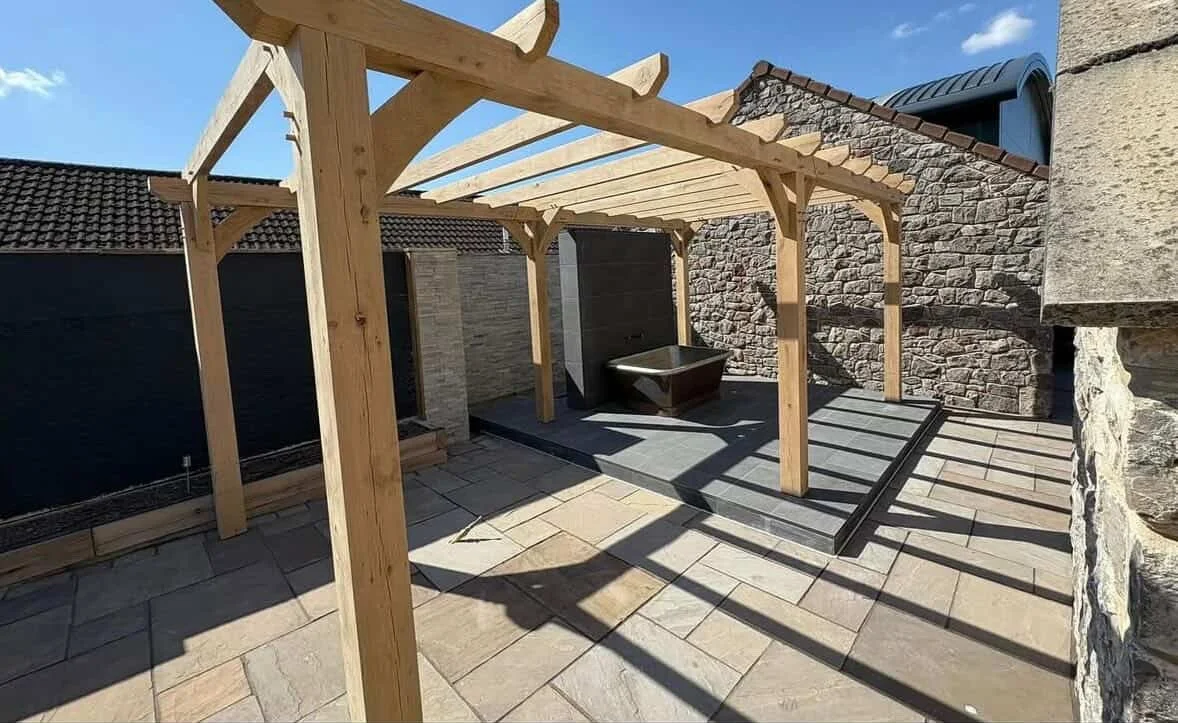 Wooden pergola construction over a patio area with stone tiles and a small raised platform, surrounded by brick and stone walls.