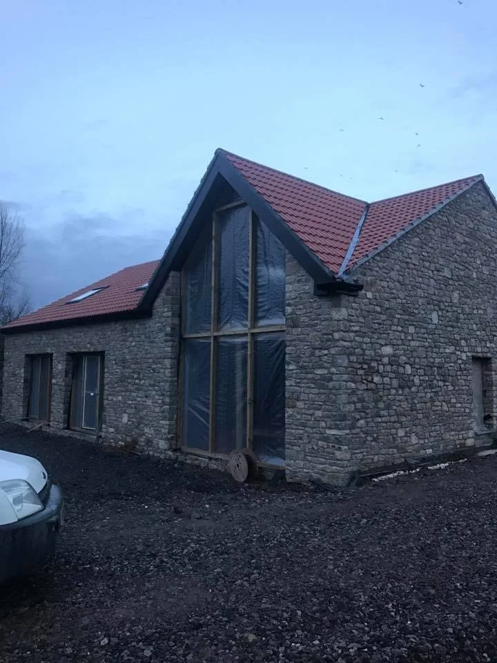 A stone house under construction with a large front window and a red tiled roof, parked cars in front, and a cloudy sky above.