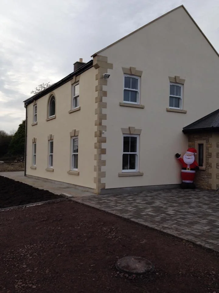 A two-story house with cream-colored exterior walls, beige brick accents around the windows, and a dark roof. There is an inflatable Santa Claus decoration next to the house entrance. The ground is partially paved with bricks, with some soil visible 