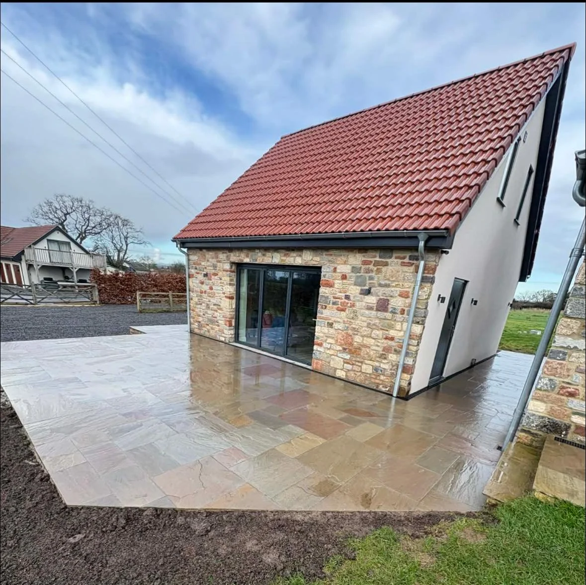 A small house with a red tiled roof, brick and white exterior walls, and large glass sliding doors, with a stamped concrete patio in the foreground, on a cloudy day.