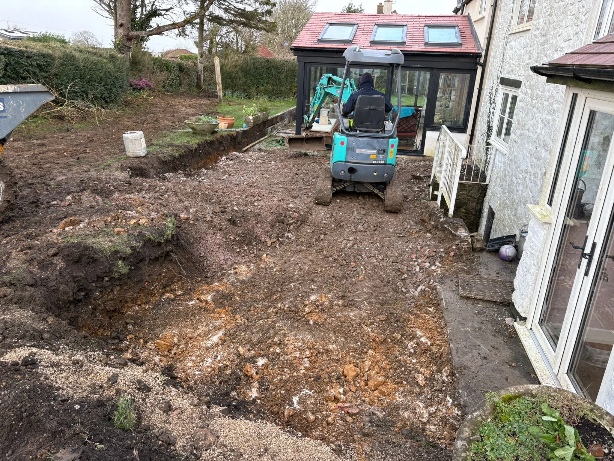 A backyard undergoing excavation with a small blue excavator and a person operating it, next to a white house with a conservatory garden room, and a garden with potted plants and a grassy area in the background.