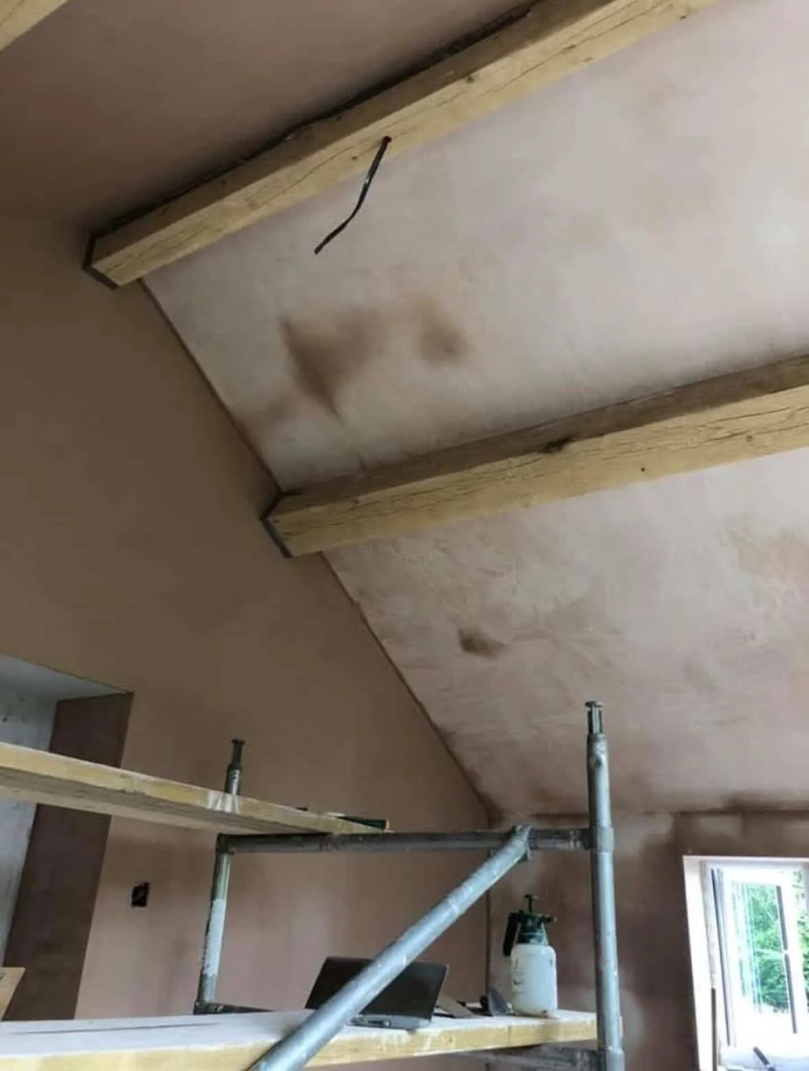 Construction scene showing a ceiling with two brown stains, supported by a wooden frame and metal scaffolding, with a window in the background.