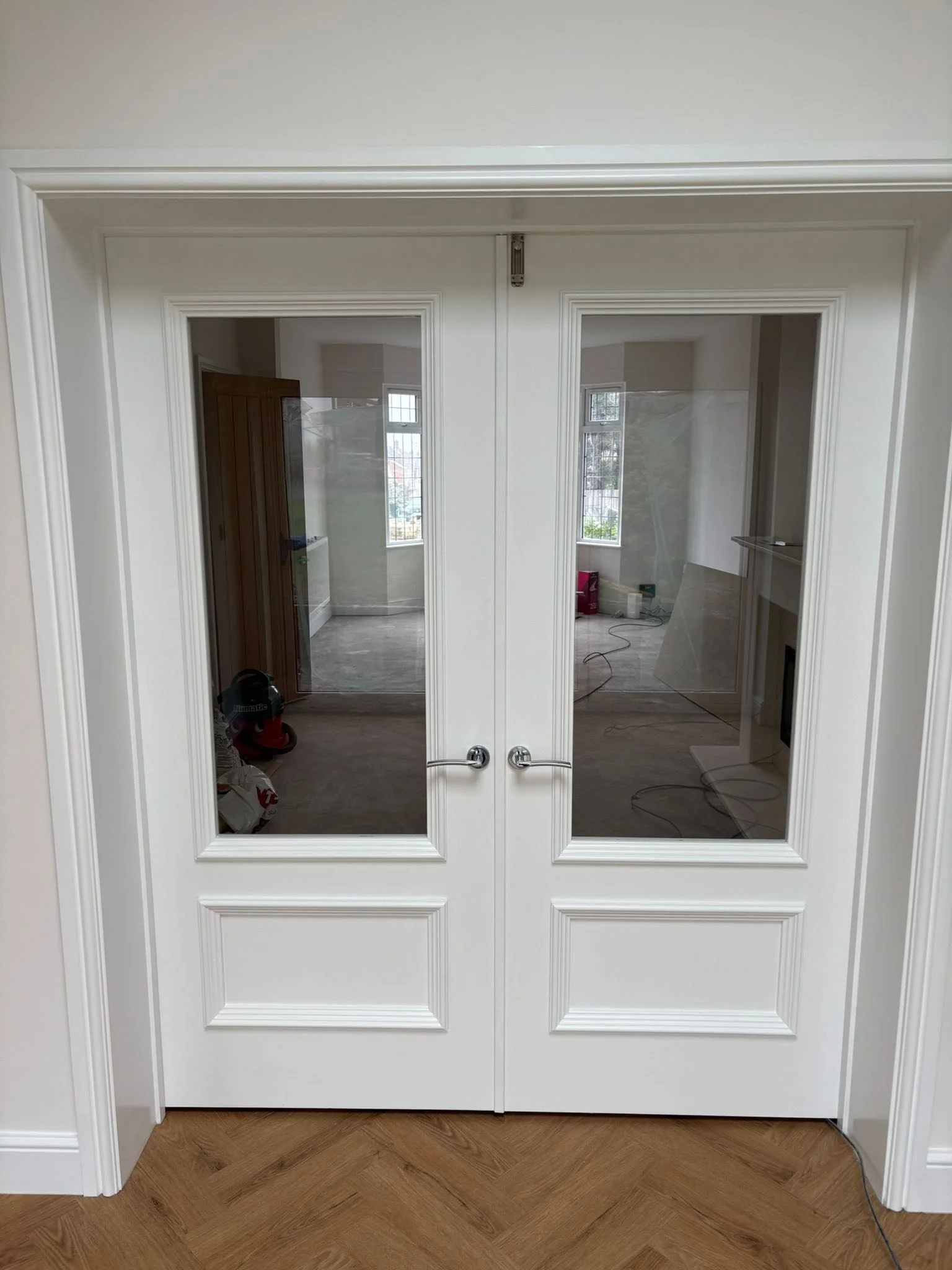 White double doors with glass panels and silver handles, opening into a room with large windows and construction tools inside.