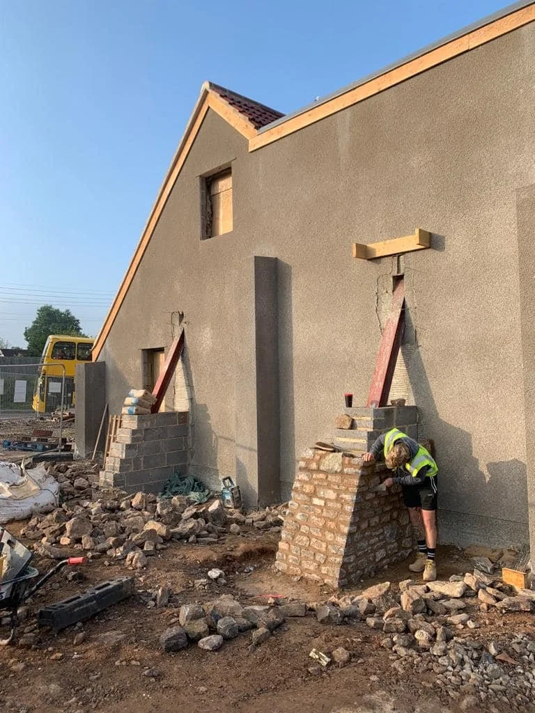 Construction worker building a brick wall in front of a gray house under construction, with tools and debris on the ground.