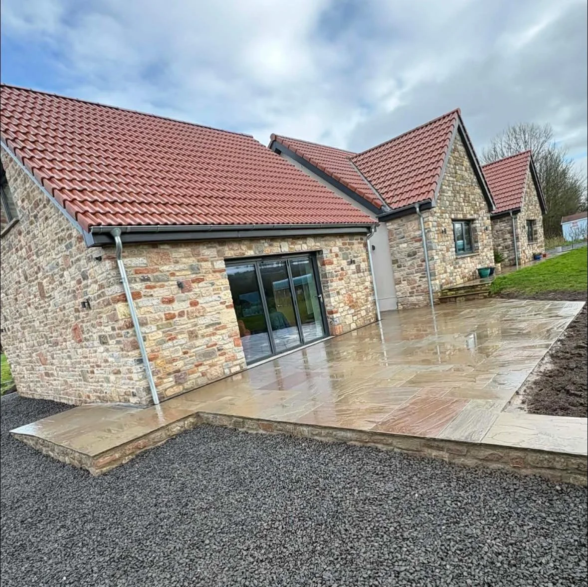 A stone house with a red tiled roof and a large glass sliding door. There is a newly paved concrete patio outside the house, with some green grass and a garden area to the right. The sky is overcast.