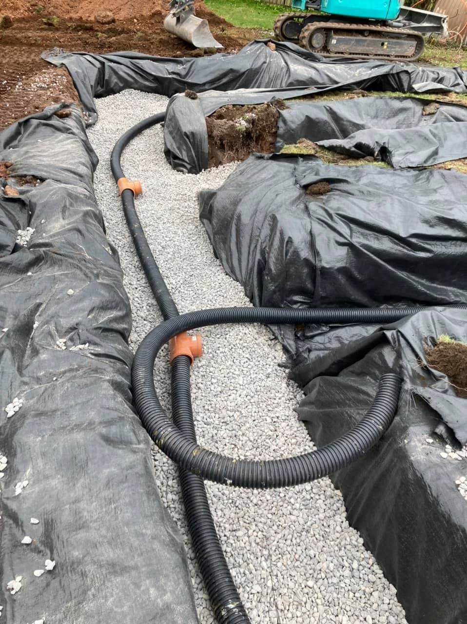 Construction site showing gravel being laid in trenches lined with black plastic sheeting, with flexible plastic pipes installed for underground utilities, and a small excavator in the background.