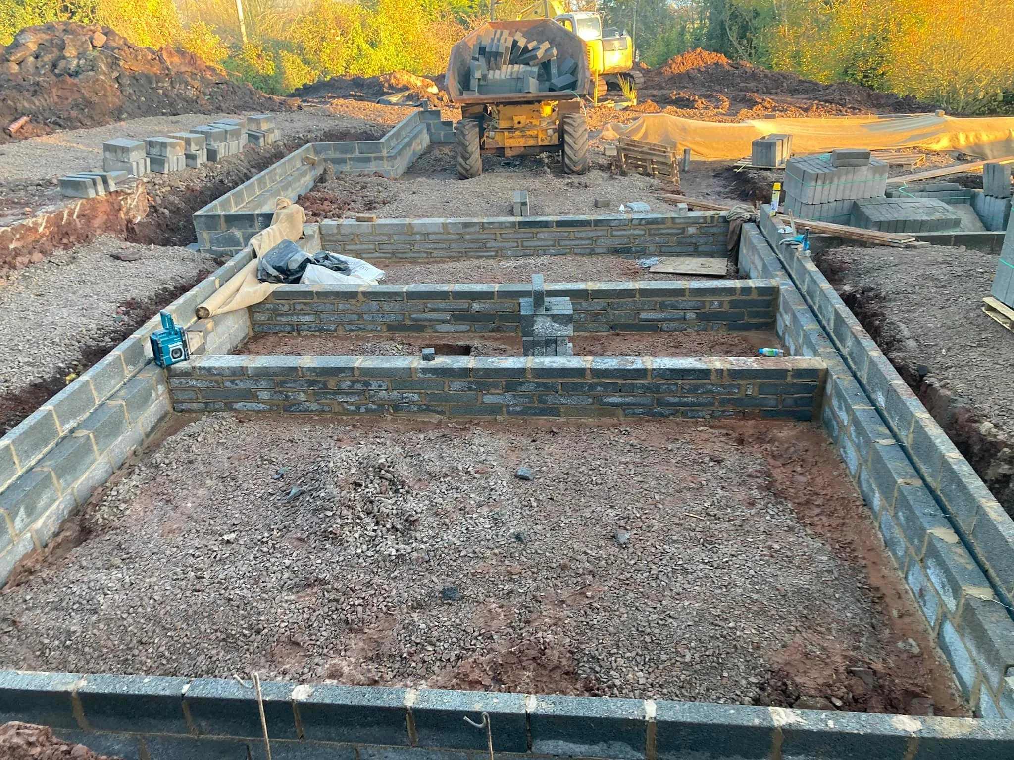 Construction site with partially built brick foundation for a building, with scattered construction materials, a construction worker, and a small excavator in the background.