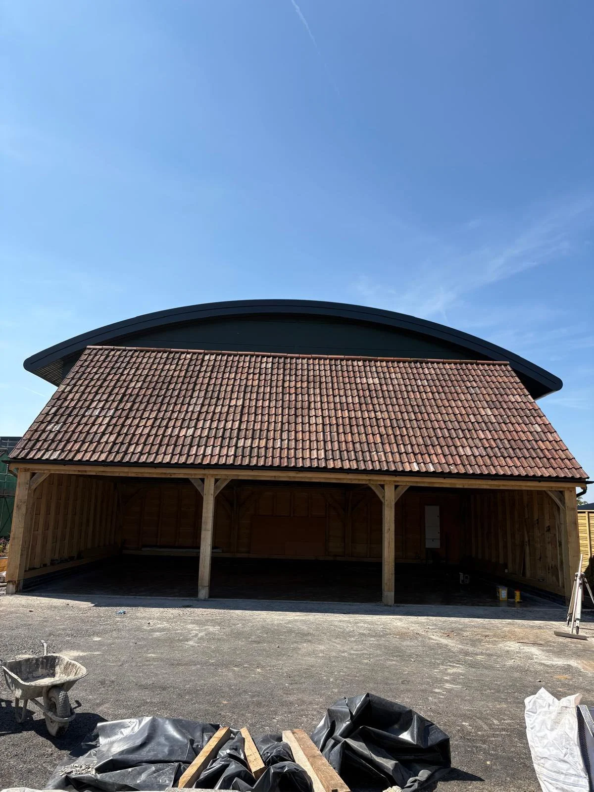 Construction of a wooden carport with a red tiled roof and black curved metal roof in the background, clear blue sky.