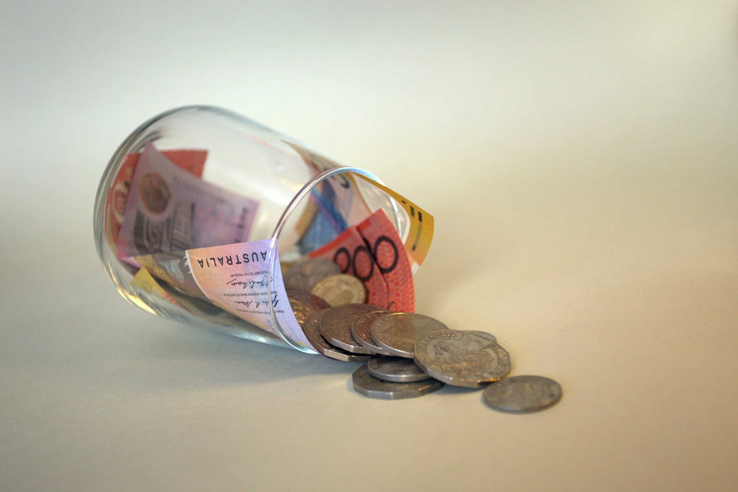 A tilted glass container spilling out various Australian dollar bills and coins onto a white surface.