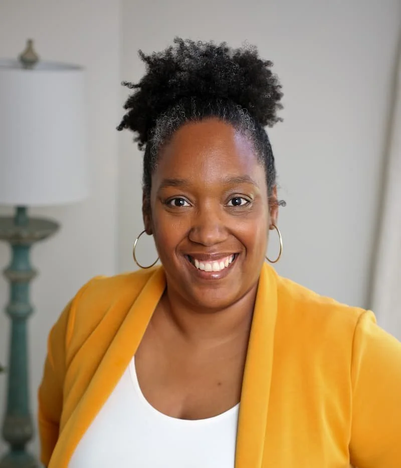 A smiling woman with curly black hair in a high puff, wearing a yellow blazer, white top, and hoop earrings, standing indoors near a lamp and light-colored curtains.