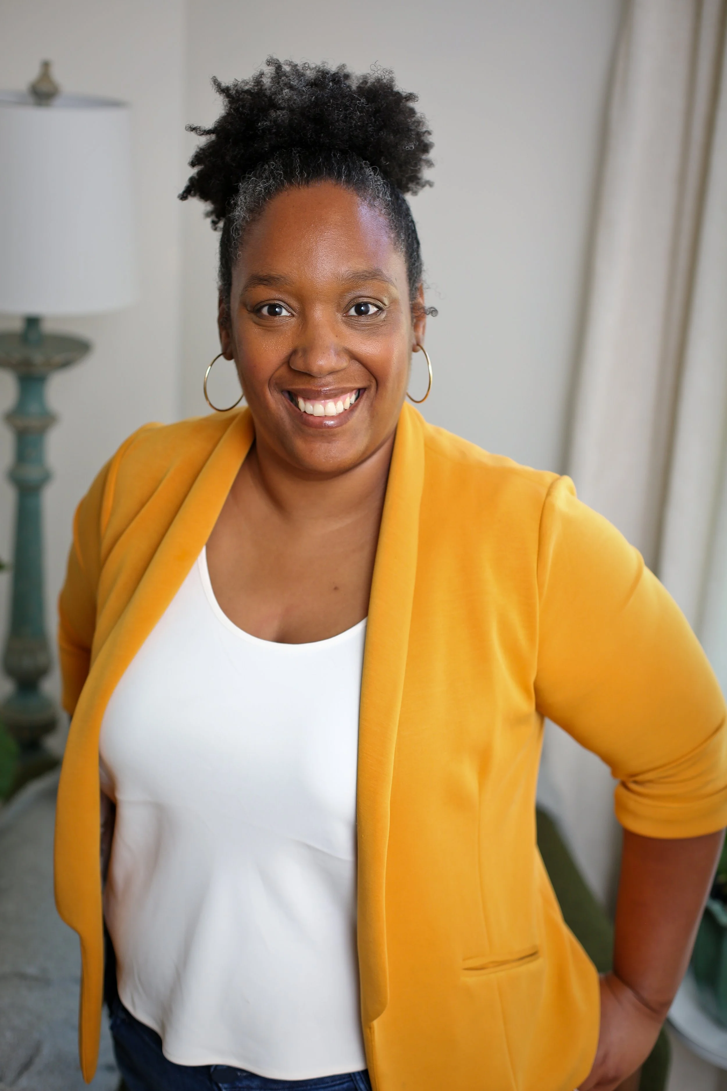 A smiling Black woman with dark natural hair styled in a puff, wearing hoop earrings, a mustard yellow blazer, and a white top, standing indoors in front of a lamp and curtains.