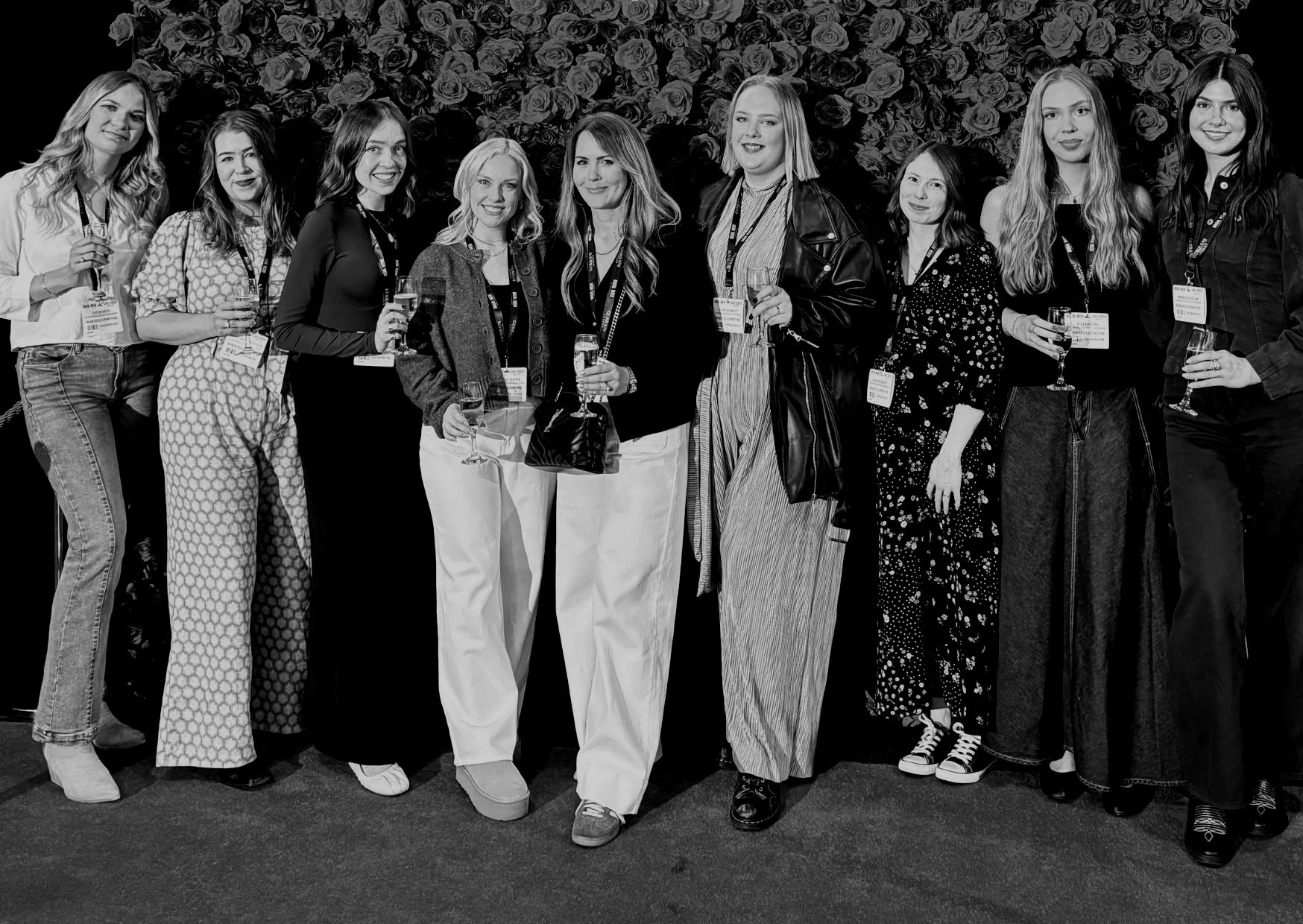 A group of eleven women standing together at an event, holding glasses of wine, with a background of roses.